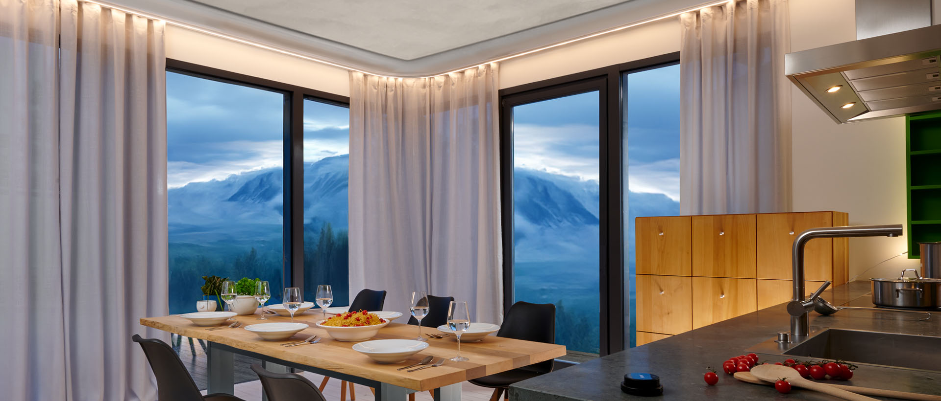 Eine offene Küche mit gedecktem Esstisch und großer Fensterfront im Hintergrund mit Blick auf Berge in der Dämmerung, der Raum wird stimmungsvoll beleuchtet durch eine gebogene Vorhangschiene mit zwei Läufen, im hinteren Lauf befinden sich helle Gardinen, im vorderen Lauf das extra warm weiße LED-Lichtband von ALUGARD.